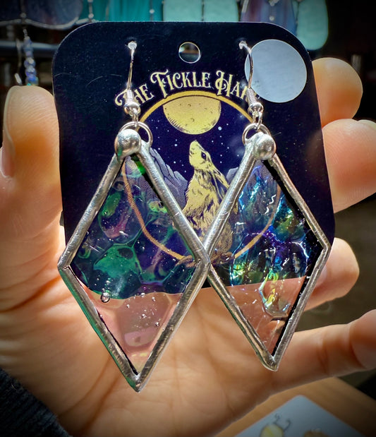 Diamond-shaped handmade stained-glass earrings with colorful iridescent glass. On a branded card held by a hand.
